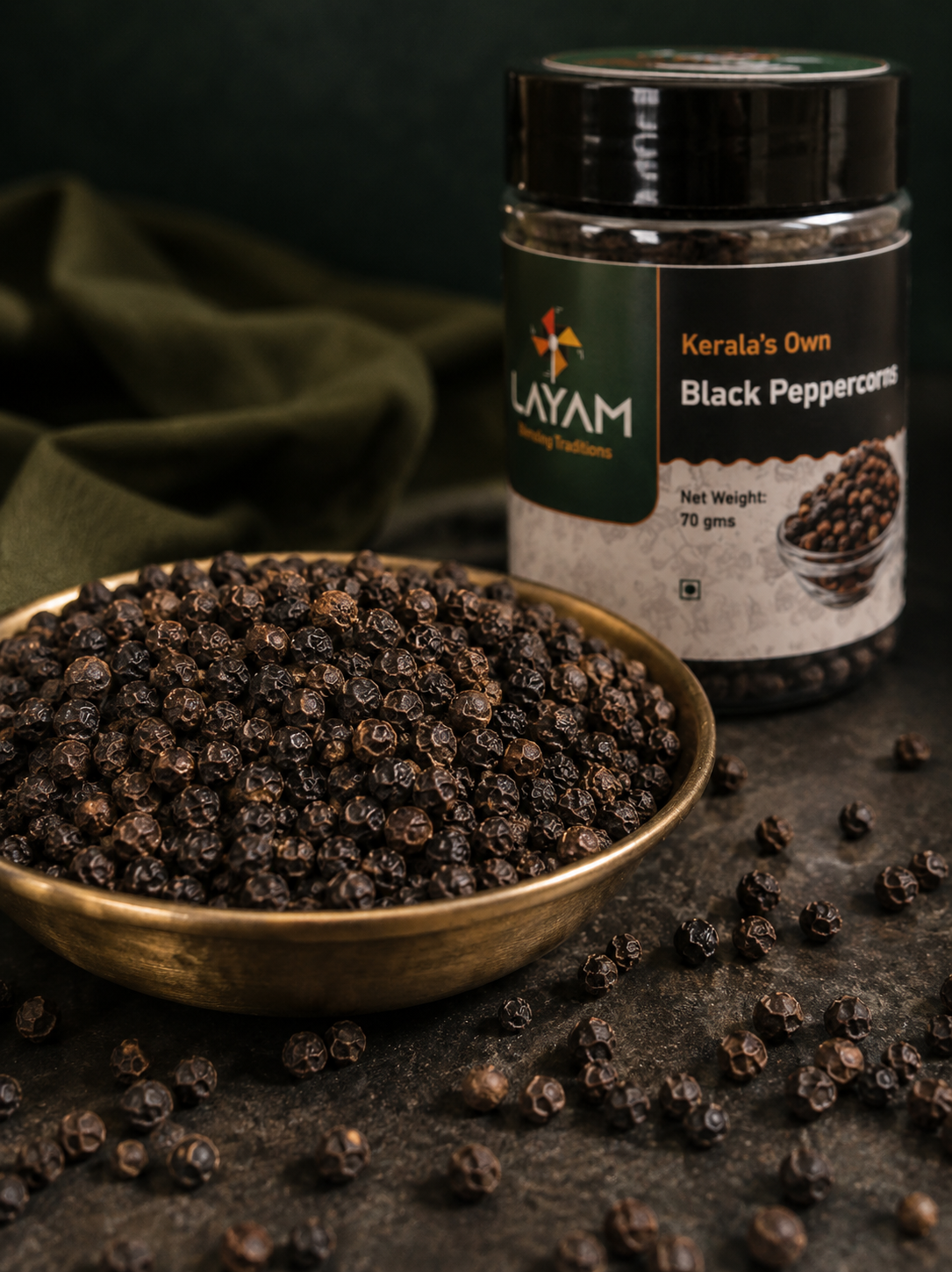 Close-up of whole sun-dried black peppercorns in a brass dish beside the LAYAM jar